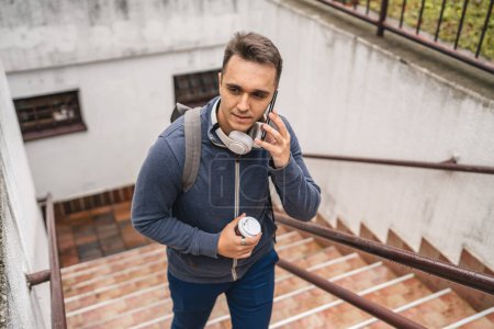 Photo for One man young adult male walking up the stairs steps in the city in day with cup of coffee and mobile phone real people tourist or student copy space use smartphone to make a call talk - Royalty Free Image
