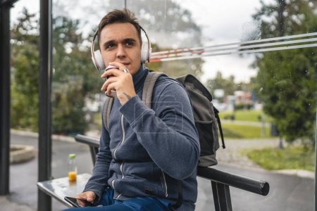 Photo for One man young adult male sit at public transport bus station waiting with headphones and mobile smart phone in winter or autumn day with backpack student or tourist city life copy space - Royalty Free Image