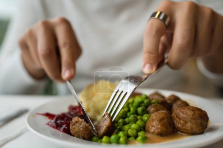 Photo for Close up on hands of unknown caucasian man sit at the table with fork and knife eat meatballs with peas and mashed potato - Royalty Free Image