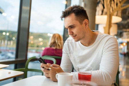 Téléchargez les photos : Un homme jeune adulte caucasien mâle assis au café ou au restaurant en utilisant un téléphone mobile faire un appel parler personnes réelles copier l'espace heureux sourire sms textos application internet en ligne - en image libre de droit