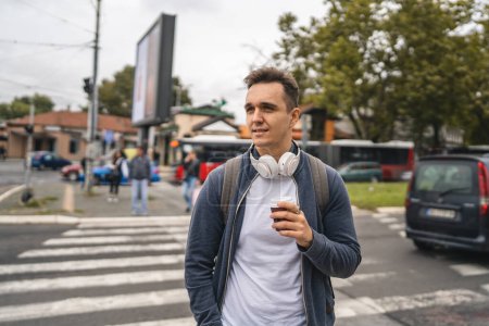 Photo for One man young adult caucasian male standing outdoor in the city waiting at crosswalk pedestrian crossing modern tourist or student in autumn day hold cup of coffee real people copy space - Royalty Free Image