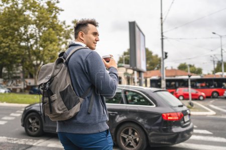 Photo for One man young adult caucasian male standing outdoor in the city waiting at crosswalk pedestrian crossing modern tourist or student in autumn day hold cup of coffee real people copy space - Royalty Free Image