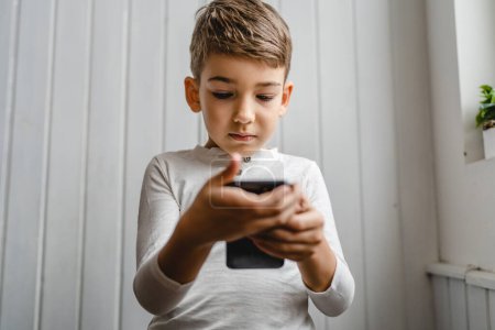 Photo for One boy caucasian child preschooler hold smartphone mobile phone at home play video games childhood and growing up technology addiction concept use smartphone app for online browsing or watch video - Royalty Free Image