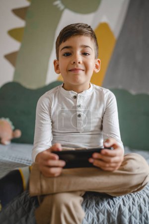 Téléchargez les photos : Un garçon caucasien enfant d'âge préscolaire tenir smartphone téléphone mobile à la maison jouer à des jeux vidéo enfance et grandir concept de dépendance à la technologie utiliser l'application smartphone pour la navigation en ligne ou regarder la vidéo - en image libre de droit