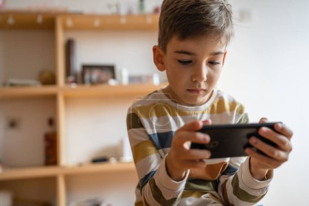 Foto de One boy caucasian child preschooler hold smartphone mobile phone at home play video games childhood and growing up technology addiction concept use smartphone app for online browsing or watch video - Imagen libre de derechos