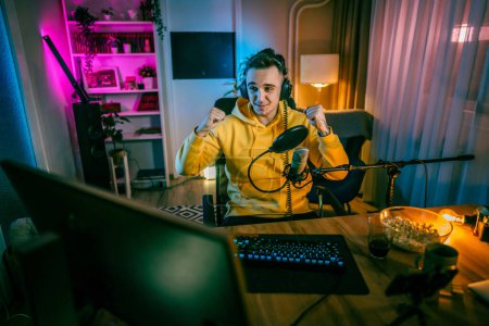Foto de Young man play video games on pc computer while streaming to social media or internet online playtrough or walktrough video male gamer having fun at home wear headphone happy winning copy space - Imagen libre de derechos
