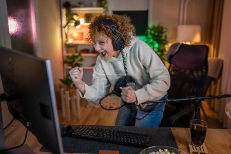 Photo for Mature woman play video games on pc computer while streaming to social media or internet online playtrough or walktrough video female gamer having fun at home wear headphone happy winning copy space - Royalty Free Image