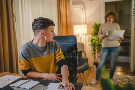 Photo for Young caucasian man teenager student study at home at the table at night or evening determinate learning prepare lesson or exam with his mother mentor or professor help real people copy space - Royalty Free Image
