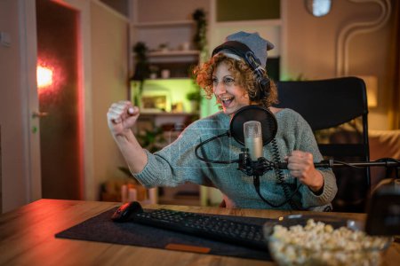 Photo for Mature woman play video games on pc computer while streaming to social media or internet online playtrough or walktrough video female gamer having fun at home wear headphone happy winning copy space - Royalty Free Image