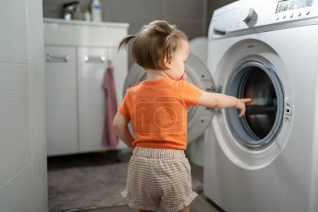 Foto de One girl small caucasian toddler child daughter standing at the washing machine in the toilet opening or closing the door examine and learn early development and growing up mischief concept copy space - Imagen libre de derechos