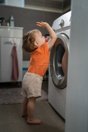 Téléchargez les photos : One girl small caucasian toddler child daughter standing at the washing machine in the toilet opening or closing the door examine and learn early development and growing up mischief concept copy space - en image libre de droit