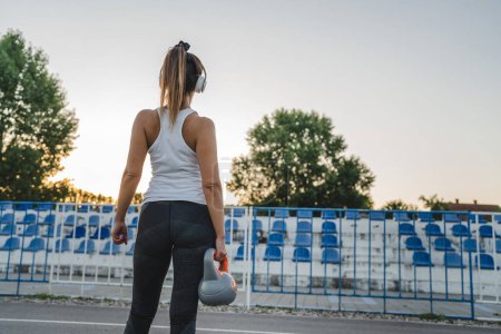 Téléchargez les photos : Back view of unknown caucasian woman female athlete holding kettlebell girya plastic weight while standing in stadium on track during training in summer evening copy space health and fitness concept - en image libre de droit