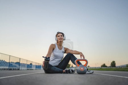 Téléchargez les photos : Une femme caucasienne athlète repos tenir supplément shaker kettlebell girya poids tandis que assis dans le stade sur la bonne voie pendant l'entraînement en été soir copie espace santé et fitness concept - en image libre de droit