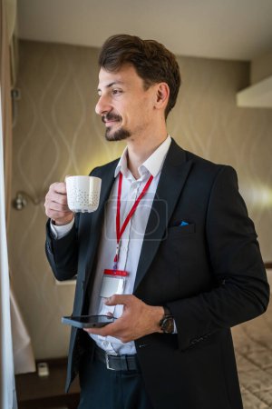 Téléchargez les photos : Un homme adulte caucasien costume masculin avec moustaches et barbe homme d'affaires dans la chambre d'hôtel tenir tasse de café avant ou après le travail en prenant un espace de copie de personnes réelles frein - en image libre de droit