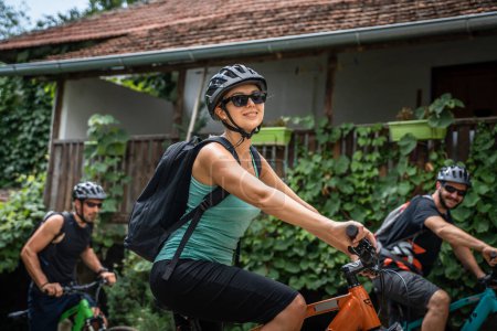 Téléchargez les photos : Group of people friends focus on One adult caucasian woman hold prepare to ride electric e-bike bicycle in sunny summer day getting ready wear protective helmet and eyeglasses real people copy space - en image libre de droit