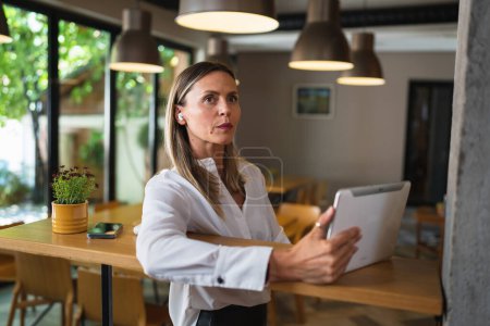 Photo for One woman adult mature caucasian female entrepreneur manager working use digital tablet while standing by the table indoor at home or at work wear white shirt copy space - Royalty Free Image
