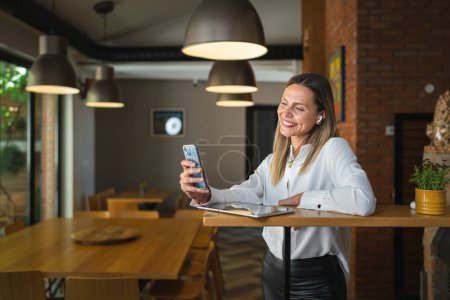 Téléchargez les photos : Une femme adulte caucasienne d'âge moyen debout au travail sourire heureux porter une chemise blanche taille vers le haut copier l'espace vue de face portrait à l'aide d'un téléphone mobile faire un appel ou envoyer sms confiant - en image libre de droit