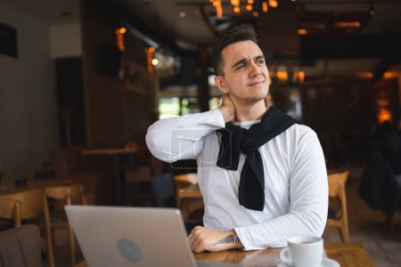 One man caucasian adult male sitting at the table at cafe restaurant using computer laptop to browse internet or work real people copy space hold neck having pain back or neck strain stretching