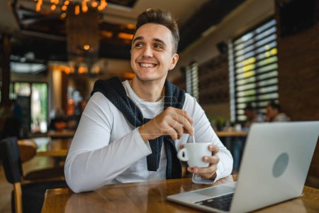 Téléchargez les photos : Un homme caucasien adulte mâle assis à la table au café restaurant à l'aide d'un ordinateur portable pour naviguer sur Internet ou travailler à distance freelance personnes réelles copier l'espace heureux sourire - en image libre de droit