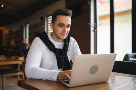 Téléchargez les photos : Un homme caucasien adulte mâle assis à la table au café restaurant à l'aide d'un ordinateur portable pour naviguer sur Internet ou travailler à distance freelance personnes réelles copier l'espace heureux sourire - en image libre de droit