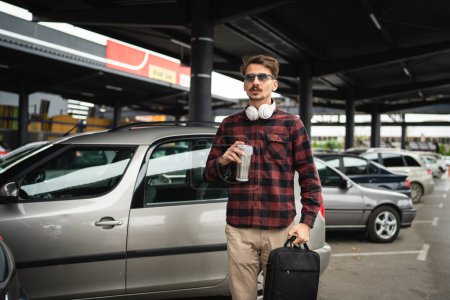 Photo for One man adult caucasian male standing by the car at the parking lot while going to work hold cup of coffee and briefcase waiting while going to or off the work real people coy space - Royalty Free Image