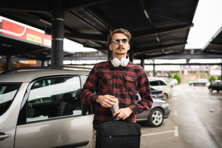 Téléchargez les photos : Un homme adulte caucasien mâle debout près de la voiture au parking tout en allant au travail tenir tasse de café et serviette d'attente tout en allant ou en dehors de l'espace de travail vraies personnes timides - en image libre de droit