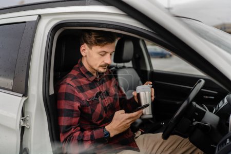 Photo for One man caucasian male with mustaches sit in the car take a brake on road-trip hold cup of coffee use mobile phone smartphone for sms texting or navigation app real people copy space - Royalty Free Image