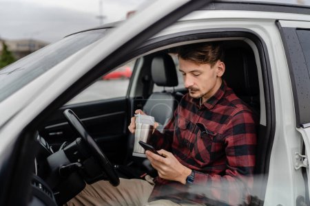 Téléchargez les photos : Un homme caucasien mâle avec moustaches assis dans la voiture prendre un frein sur la route tenir tasse de café utiliser téléphone portable smartphone pour SMS textos ou application de navigation personnes réelles copier l'espace - en image libre de droit
