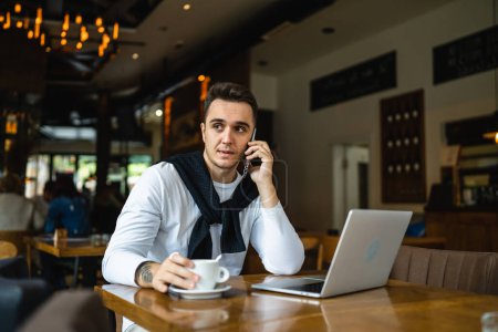 Photo for One man sitting at the table at cafe or restaurant wear casual using mobile phone to make a call talking communication while hold a cup of coffee real people copy space - Royalty Free Image