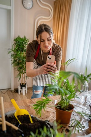 Foto de Una mujer joven generación z adulto caucásico hembra cuidar de sus plantas en casa fotografiando maceta con su teléfono inteligente móvil enviar fotos a las redes sociales o como mensaje persona real - Imagen libre de derechos