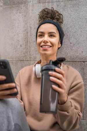 Photo for One young woman with headphones hold supplement shaker bottle and smartphone mobile phone sporty caucasian female sit take a brake for training generation z healthy lifestyle concept copy space - Royalty Free Image