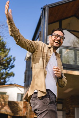 Foto de Un hombre caucásico adulto varón pie al aire libre delante de la pequeña casa moderna anfitrión o vendedor con las manos gesto invitar en la invitación gesto sonrisa feliz en el espacio de copia de día soleado - Imagen libre de derechos