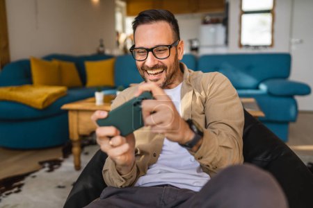 Photo for One adult caucasian man sit at home happy smile play video games leisure activity having fun hold mobile phone smartphone have fun copy space wear eyeglasses and shirt - Royalty Free Image