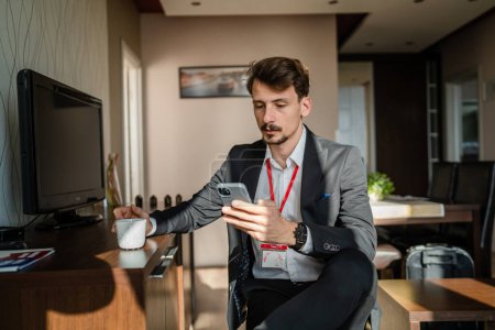 Photo for One man caucasian male adult businessman sitting at hotel room while take a brake from work use smarphone mobile phone for text messages or browse internet online hold cup of coffee in pause happy - Royalty Free Image