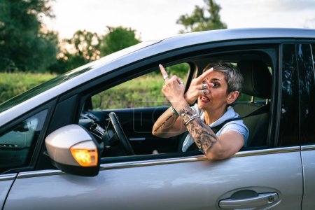 Téléchargez les photos : One woman senior mature female driver showing middle finger trough the window of the car mad angry furious pissed shows a negative gesture to the camera emotionally provocation and rude attitude - en image libre de droit