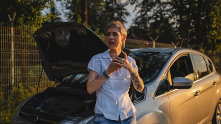 Téléchargez les photos : One woman mature female standing on the road in the evening sunset by the broken vehicle car automobile failed engine open hood making a phone call for help roadside assistance towing service concept - en image libre de droit