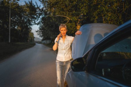 Photo for One woman mature female standing on the road in the evening sunset by the broken vehicle car automobile failed engine open hood making a phone call for help roadside assistance towing service concept - Royalty Free Image