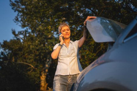 Photo for One woman mature female standing on the road in the evening sunset by the broken vehicle car automobile failed engine open hood making a phone call for help roadside assistance towing service concept - Royalty Free Image