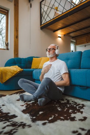 Téléchargez les photos : Un homme vieux caucasien yeux masculins fermés pour la formation guidée yoga ou méditation tout en étant assis à la maison avec casque pratique d'auto-soins de vraies personnes bien-être concept de paix intérieure et d'équilibre - en image libre de droit