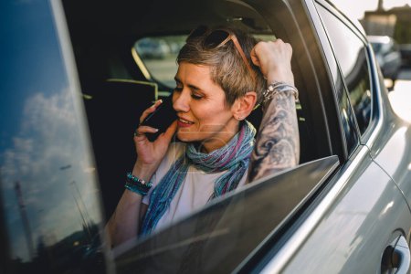 Photo for One mature woman caucasian female sitting on the back seat of the car working on laptop computer make a phone call talk in summer day with short gray hair modern on road wearing casual copy space - Royalty Free Image