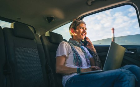 Photo for One mature woman caucasian female sitting on the back seat of the car working on laptop computer make a phone call talk in summer day with short gray hair modern on road wearing casual copy space - Royalty Free Image