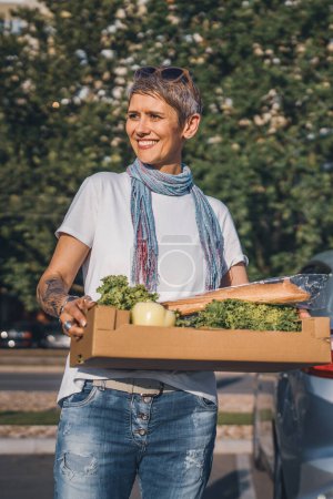Téléchargez les photos : Une femme mature femme caucasienne debout dans le parking bu voiture en face de supermarché épicerie tenant boîte avec de la nourriture et des légumes dans la journée ensoleillée sourire heureux saine manger concept végétalien - en image libre de droit