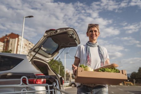 Foto de Una mujer madura caucásica hembra de pie en el estacionamiento bu coche en frente de supermercado tienda de comestibles caja de espera con alimentos y verduras en el día soleado sonrisa feliz comer sano concepto vegano - Imagen libre de derechos