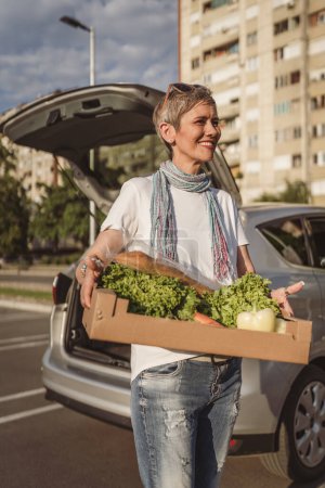 Foto de Una mujer madura caucásica hembra de pie en el estacionamiento bu coche en frente de supermercado tienda de comestibles caja de espera con alimentos y verduras en el día soleado sonrisa feliz comer sano concepto vegano - Imagen libre de derechos