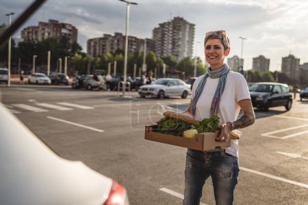 Photo for One woman mature caucasian female standing in the parking lot bu car in front of supermarket grocery store holding box with food and vegetables in sunny day happy smile healthy eating vegan concept - Royalty Free Image