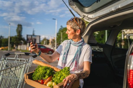 Téléchargez les photos : Une femme mature caucasienne assise dans le coffre arrière de sa voiture sur le parking du supermarché centre commercial ou épicerie avec des légumes nourriture dans la boîte les mettre dans le véhicule - en image libre de droit