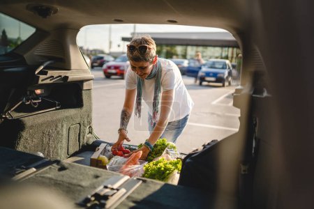 Téléchargez les photos : Une femme caucasienne mature se tient au coffre arrière de sa voiture sur le parking du supermarché centre commercial ou épicerie avec des légumes nourriture dans la boîte de les mettre dans le véhicule - en image libre de droit