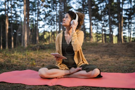 Photo for One woman young adult caucasian female sitting on the yoga mat alone in the park or forest in nature holding digital tablet and adjusting headphones preparing for online guided meditation self care - Royalty Free Image