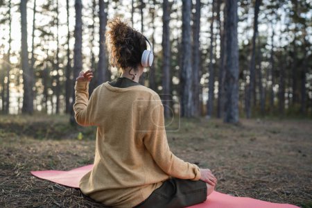 Téléchargez les photos : Une femme jeune femme caucasienne adulte assise seule dans le parc ou dans la forêt dans la nature avec un casque pour la méditation guidée en ligne équilibre mental et pratiques de manifestation concept - en image libre de droit