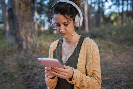 Photo for One woman young adult caucasian female sitting alone in the park or forest in nature holding digital tablet and headphones preparing for guided meditation self-care manifestation practices concept - Royalty Free Image